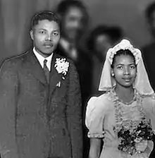A black and white photograph of a black man and a black woman, standing side by side. He is wearing a suit, she is wearing a white dress.