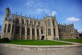 A photograph of a huge stone chapel, with tall stained glass windows and stone detailing. A patch of green grass can be seen in the foreground with several people walking by the door of the chapel.